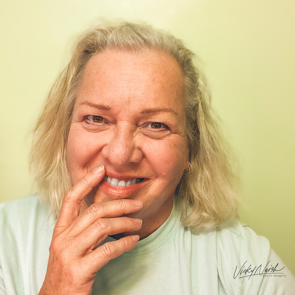 portrait of smiling blonde woman in pale green shirt on pale yellow background  woman has hand gently place by her chin and mouth and is looking straight at the camera