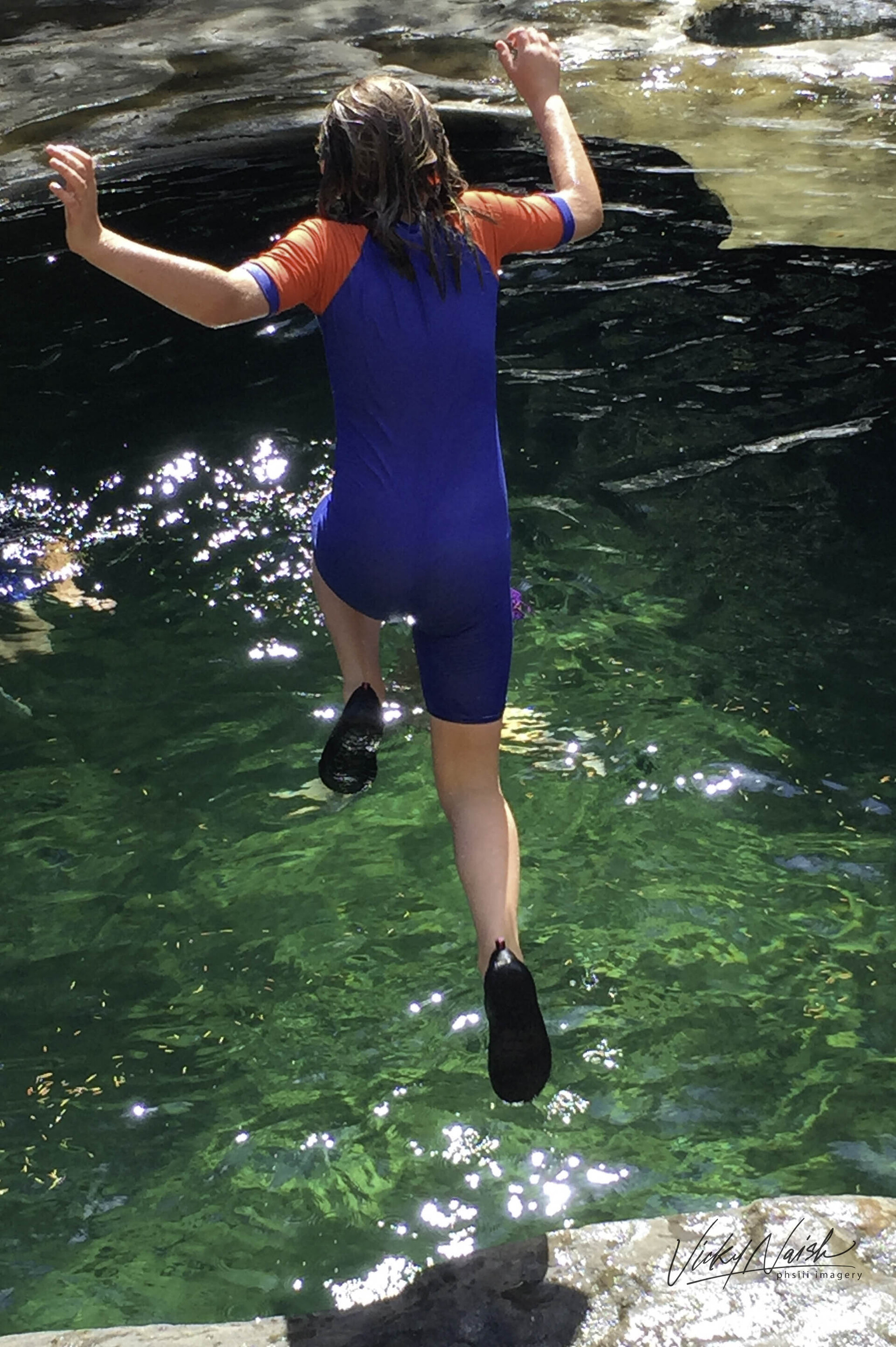 girl in UV sunsuit swimsuit and water shoes jumping off a rock cliff into glacier fed river
